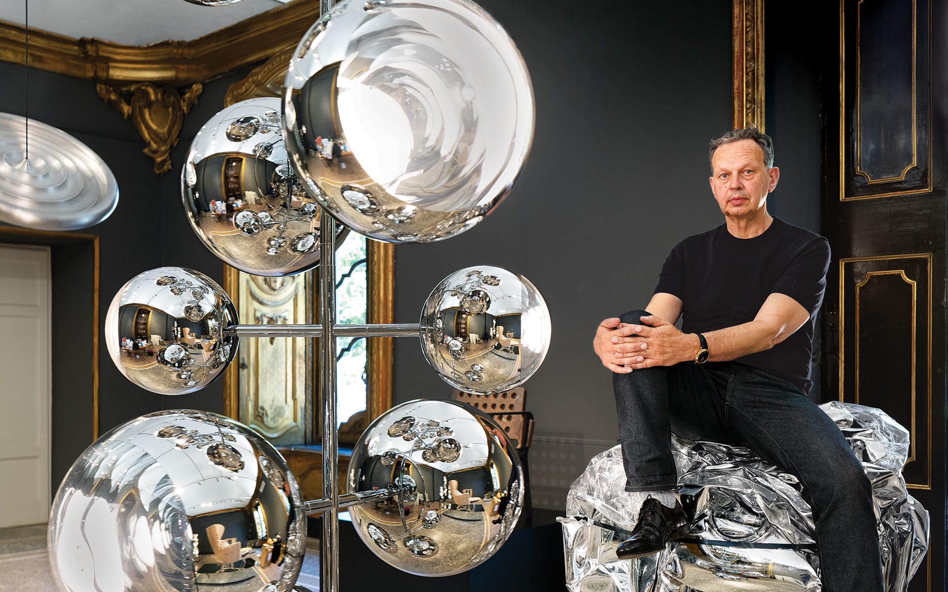 Man sitting on metallic structure surrounded by reflective spherical sculptures in elegant room with dark walls and ornate details.
