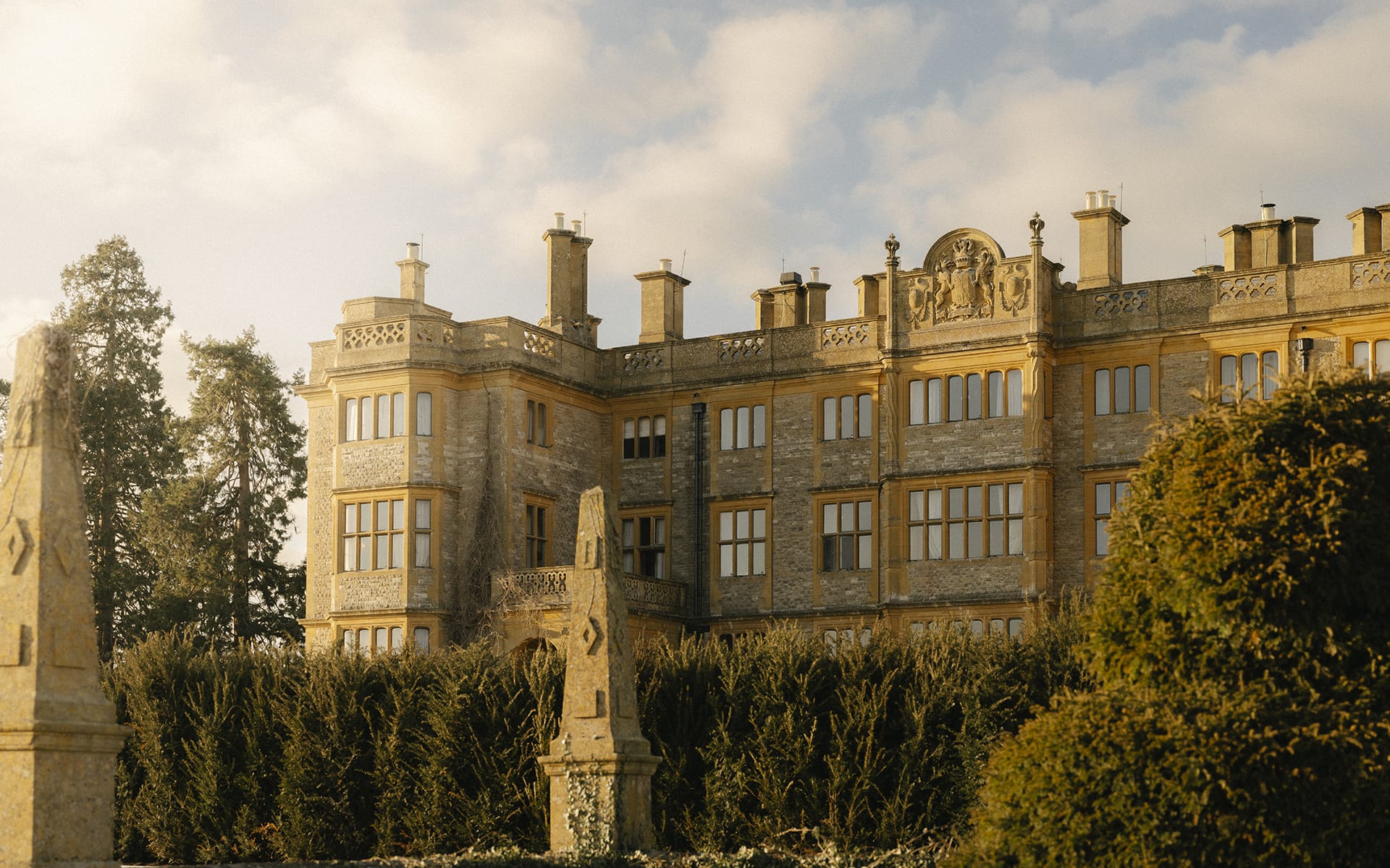 Historic stone mansion with ornate architecture surrounded by trees under a partly cloudy sky.
