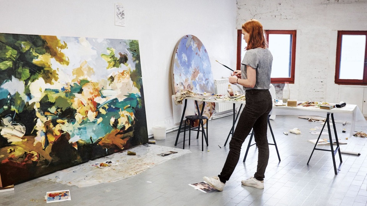 Artist standing in studio with large colorful paintings on canvas, surrounded by art supplies and photographs.