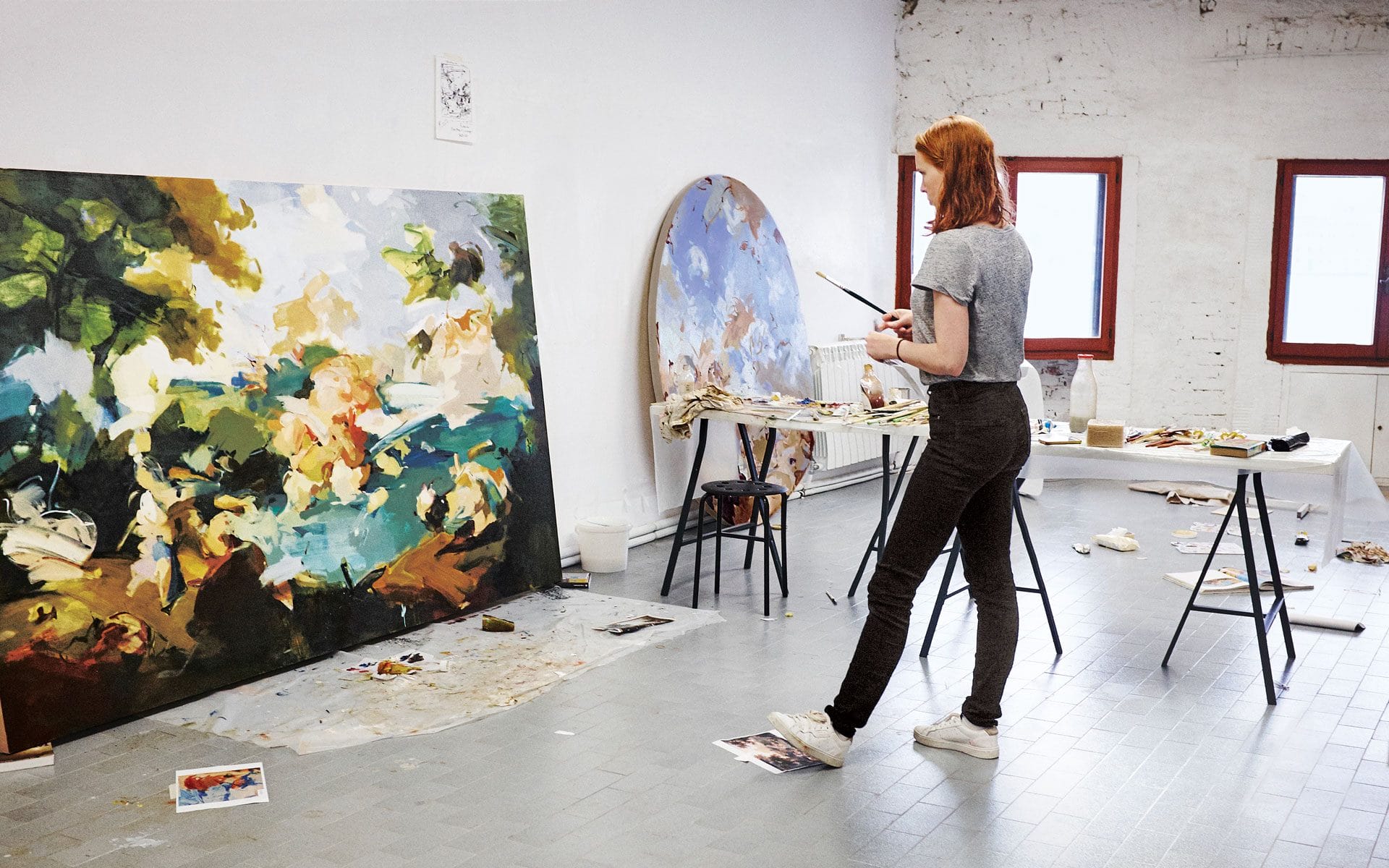 Artist standing in studio with large colorful paintings on canvas, surrounded by art supplies and photographs.