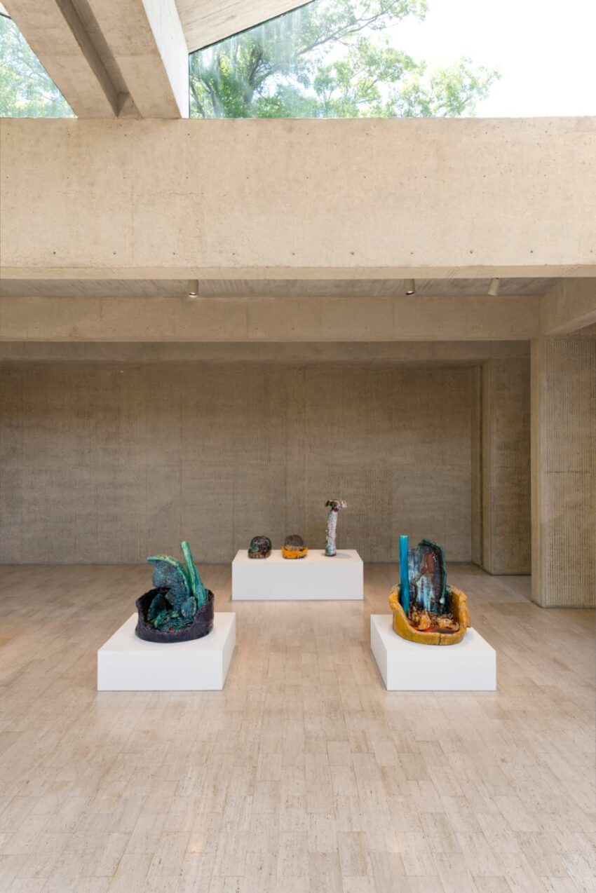 Art installation in a modern gallery with four colorful sculptures on white pedestals and a concrete ceiling with skylight.