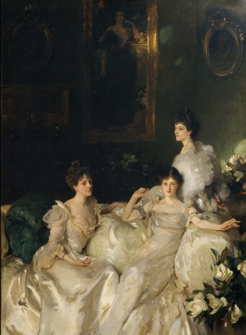 Three women in elegant white dresses sitting on a sofa in a dimly lit, ornate room with floral arrangements and framed portraits.