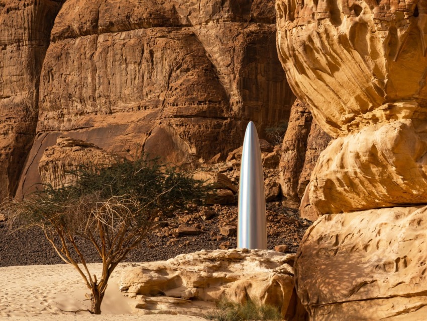 Tall silver sculpture set against rugged desert rock formations and sandy landscape.