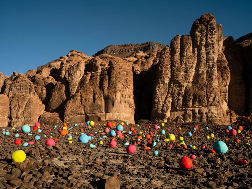 Colorful spheres scattered on rocky desert landscape with towering cliffs under a clear blue sky.