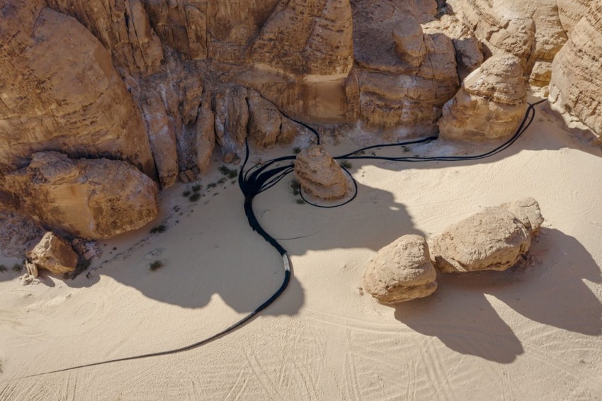 Aerial view of a desert landscape with winding black tubes or cables among large rock formations and sandy ground.