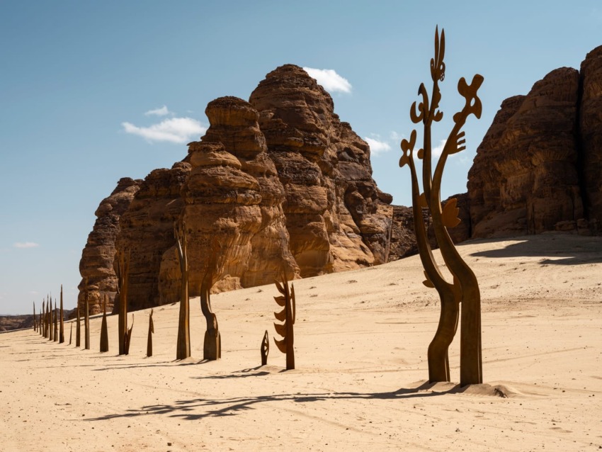 Artwork of tall metal sculptures in a desert landscape with rocky formations in the background under a blue sky.