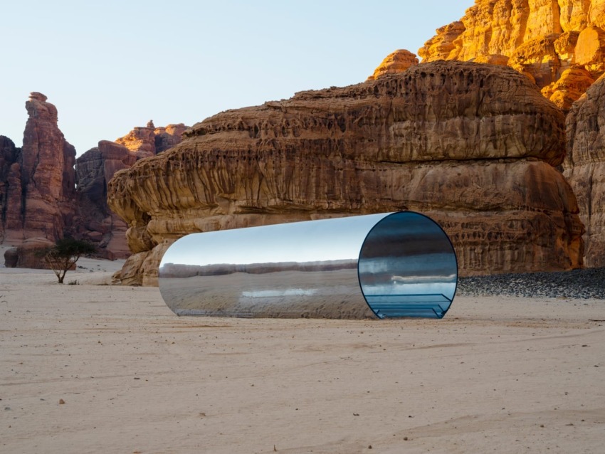 Mirrored cylindrical art installation in a desert landscape with rocky cliffs in the background.
