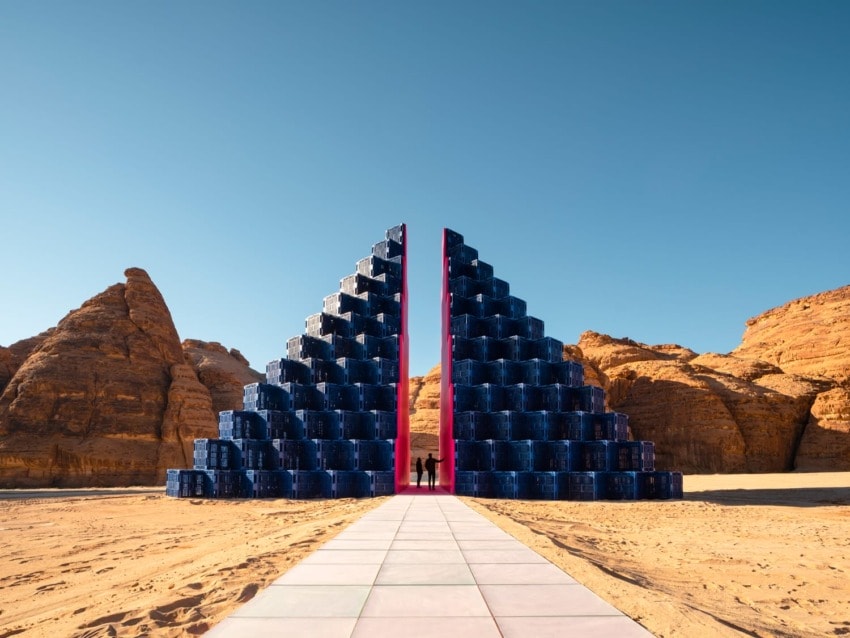 Modern art installation with blue barrels forming a pyramid split by a vibrant pink line, set in a desert landscape.