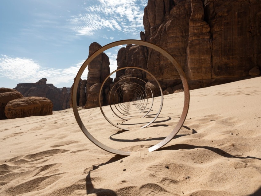 Artistic metal rings align in desert sand with rocky cliffs and a partly cloudy sky in the background.