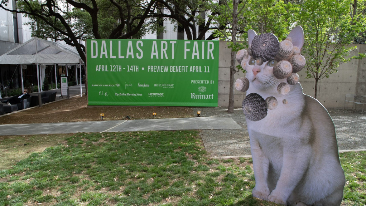 Sign for Dallas Art Fair April 12-14 with cat sculpture adorned with spherical objects in outdoor setting.