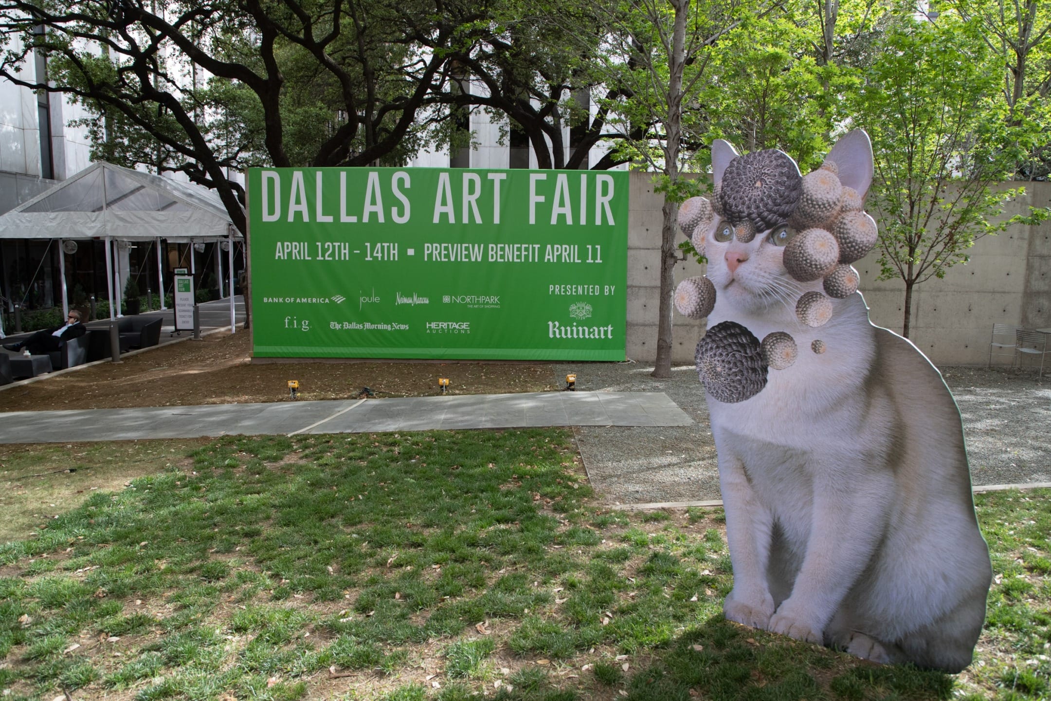 Sign for Dallas Art Fair April 12-14 with cat sculpture adorned with spherical objects in outdoor setting.
