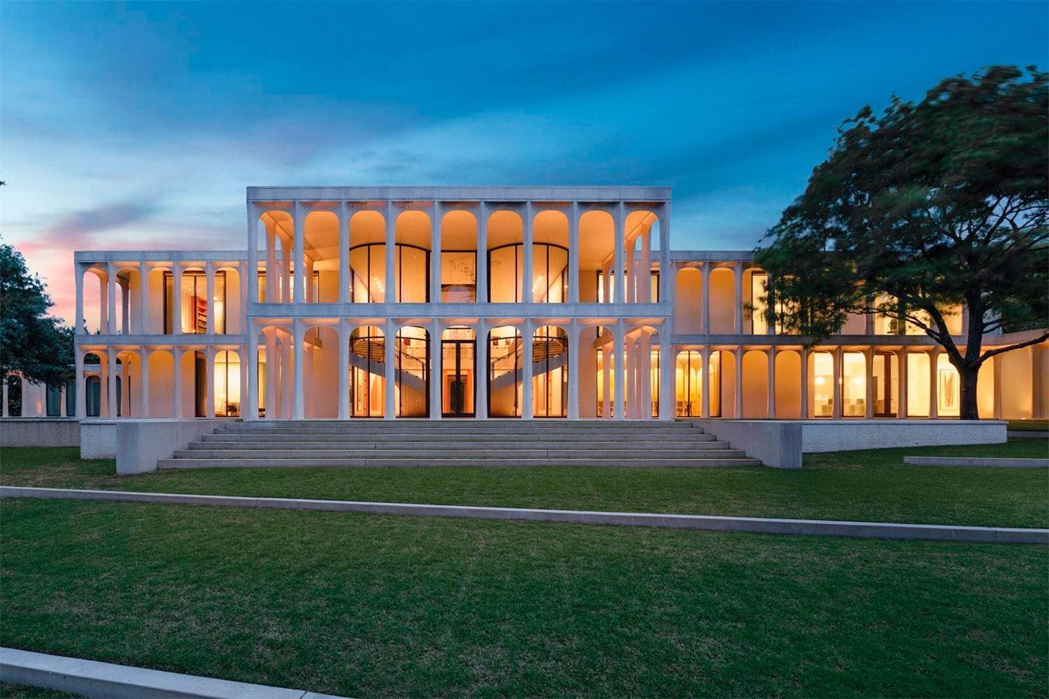 Modern building with illuminated arches and windows, surrounded by a grassy lawn and tree, during twilight.