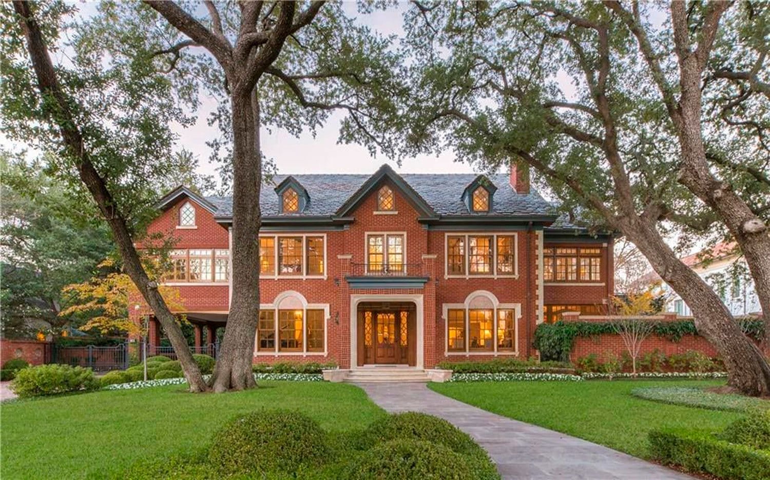 Stately brick mansion with large windows, surrounded by lush green lawn and trees, set against a clear sky.