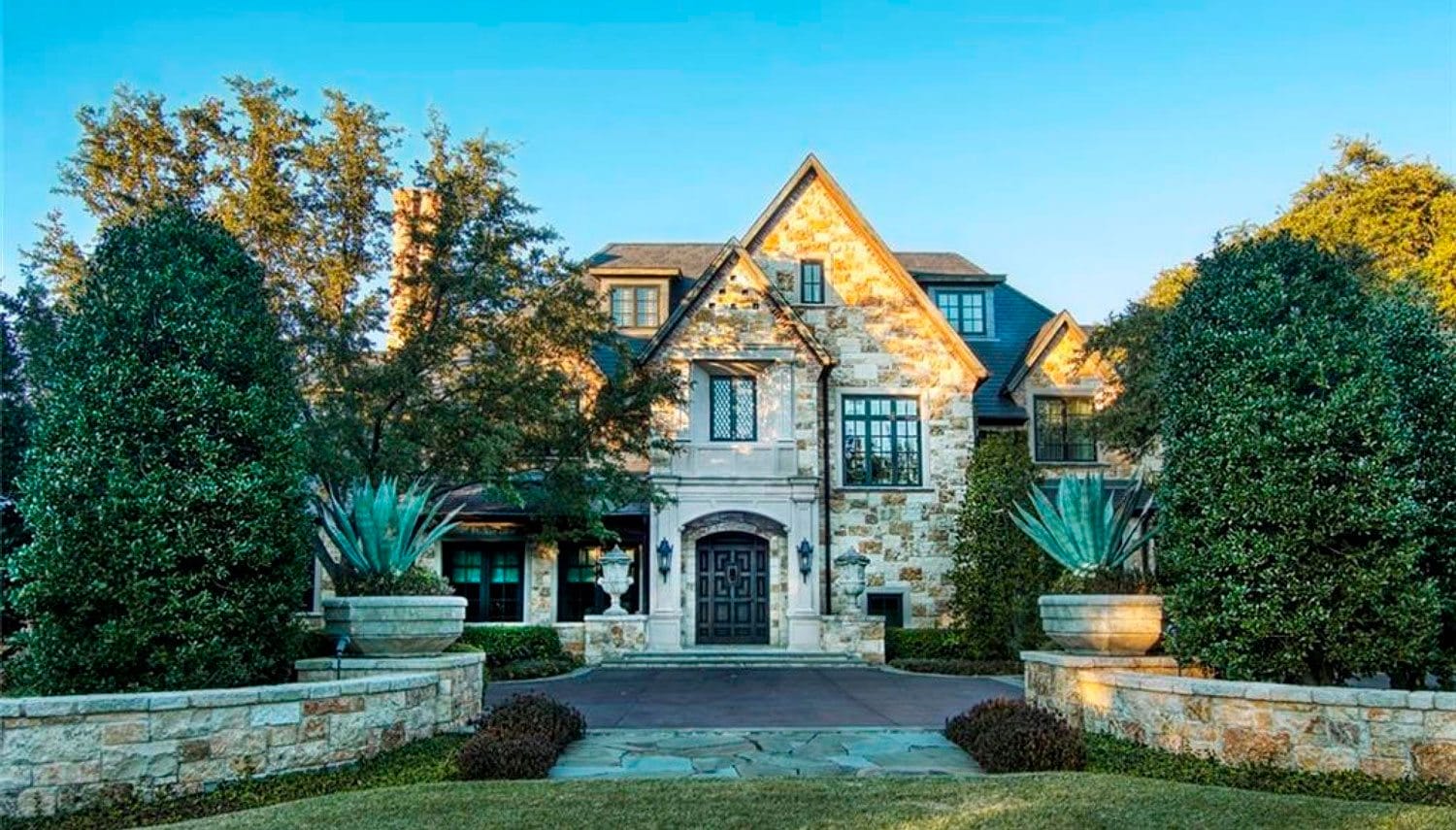 Large stone house with lush landscaping, symmetrical design, tall trees, and a paved driveway on a sunny day.