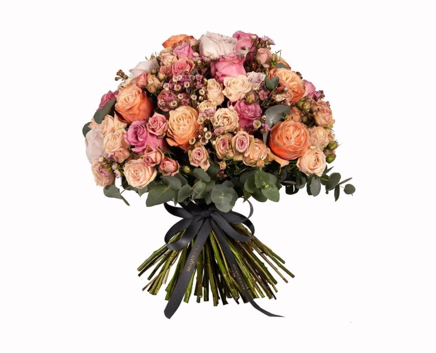 Bouquet of mixed pink, orange, and peach roses with green leaves wrapped in a black ribbon.