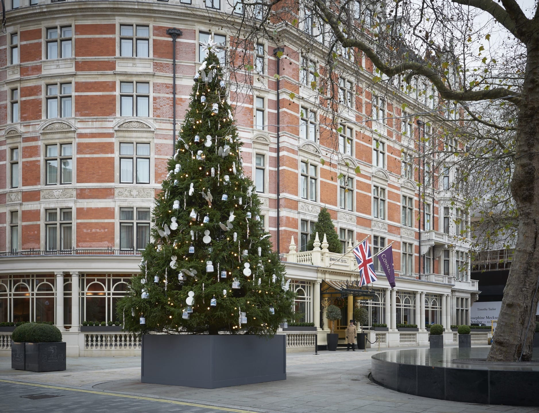 Large decorated Christmas tree in front of a red brick building with multiple windows and Union Jack flag.