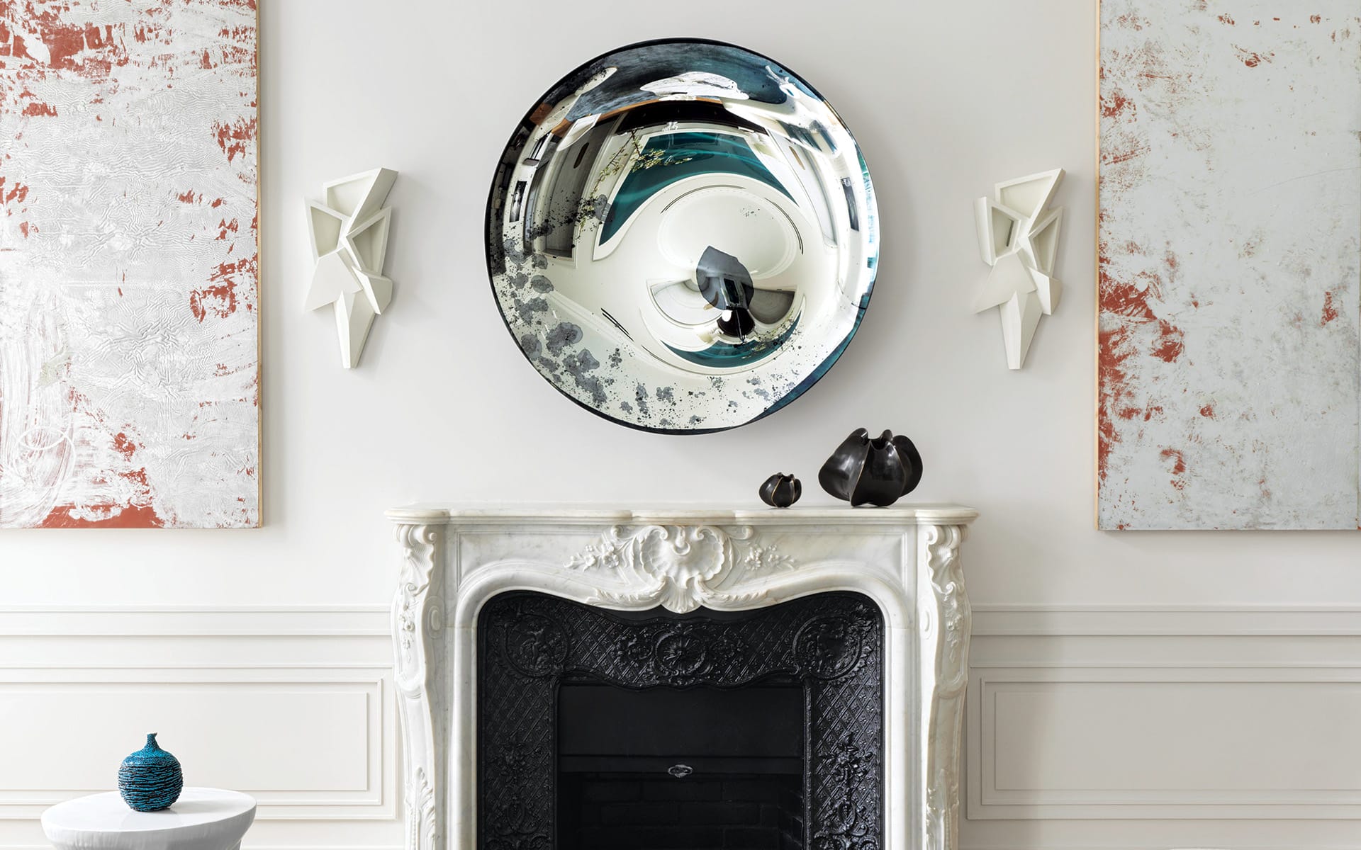 Elegant living room with a white marble fireplace, decorative round mirror, abstract art, and modern sculptures.