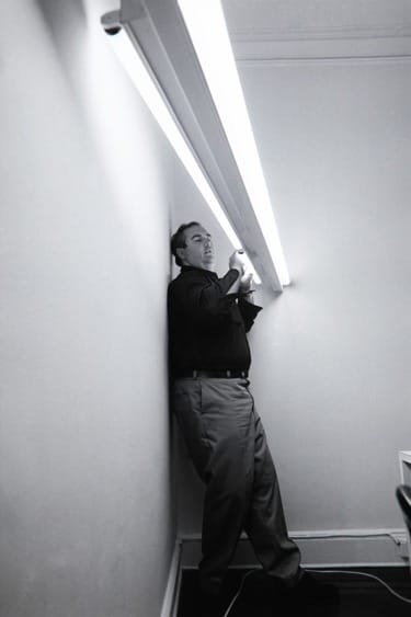 Man standing against a wall adjusting a long fluorescent light tube in a room with white walls and dark flooring.
