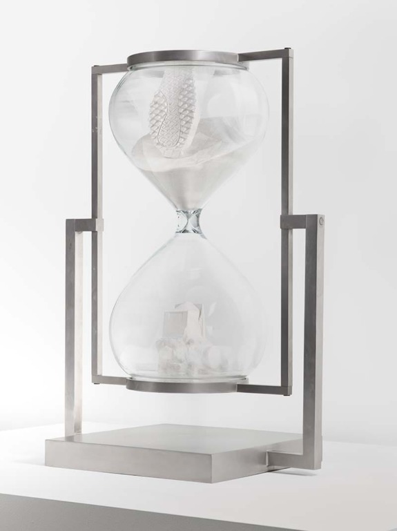 Modern hourglass sculpture with metal frame and intricate internal designs on a white background.