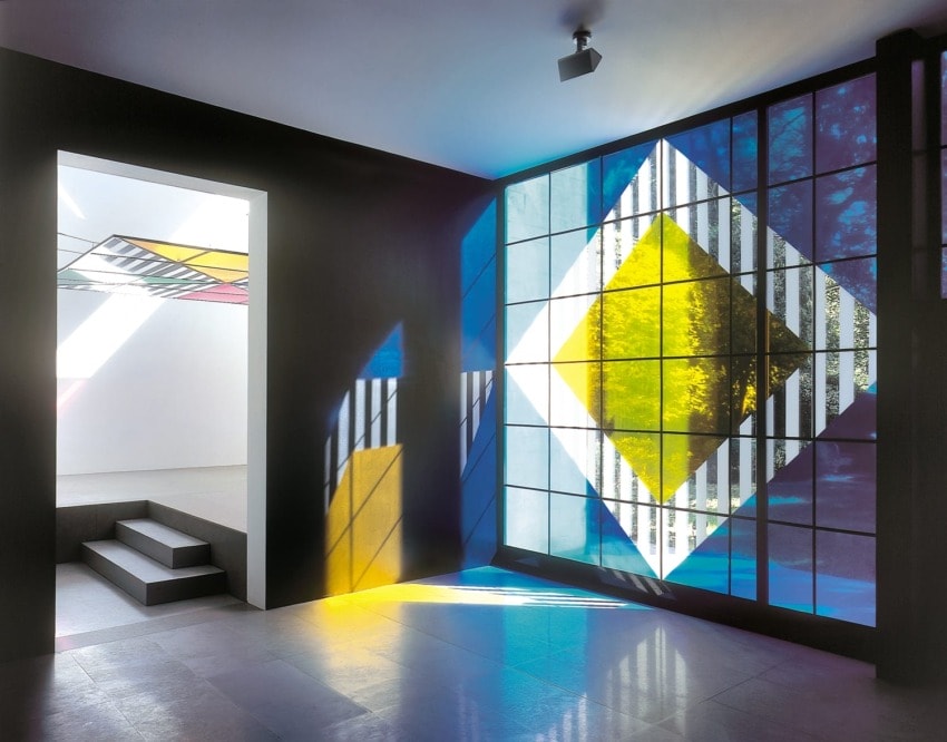 Colorful geometric patterns casting shadows in a modern interior, with stairs leading to another room filled with light.