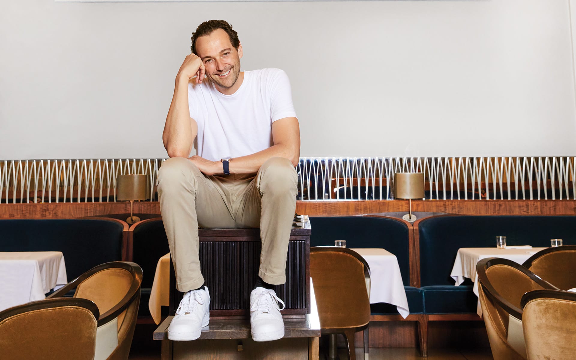 Man smiling while sitting on a box in a stylish restaurant with empty tables and modern decor.