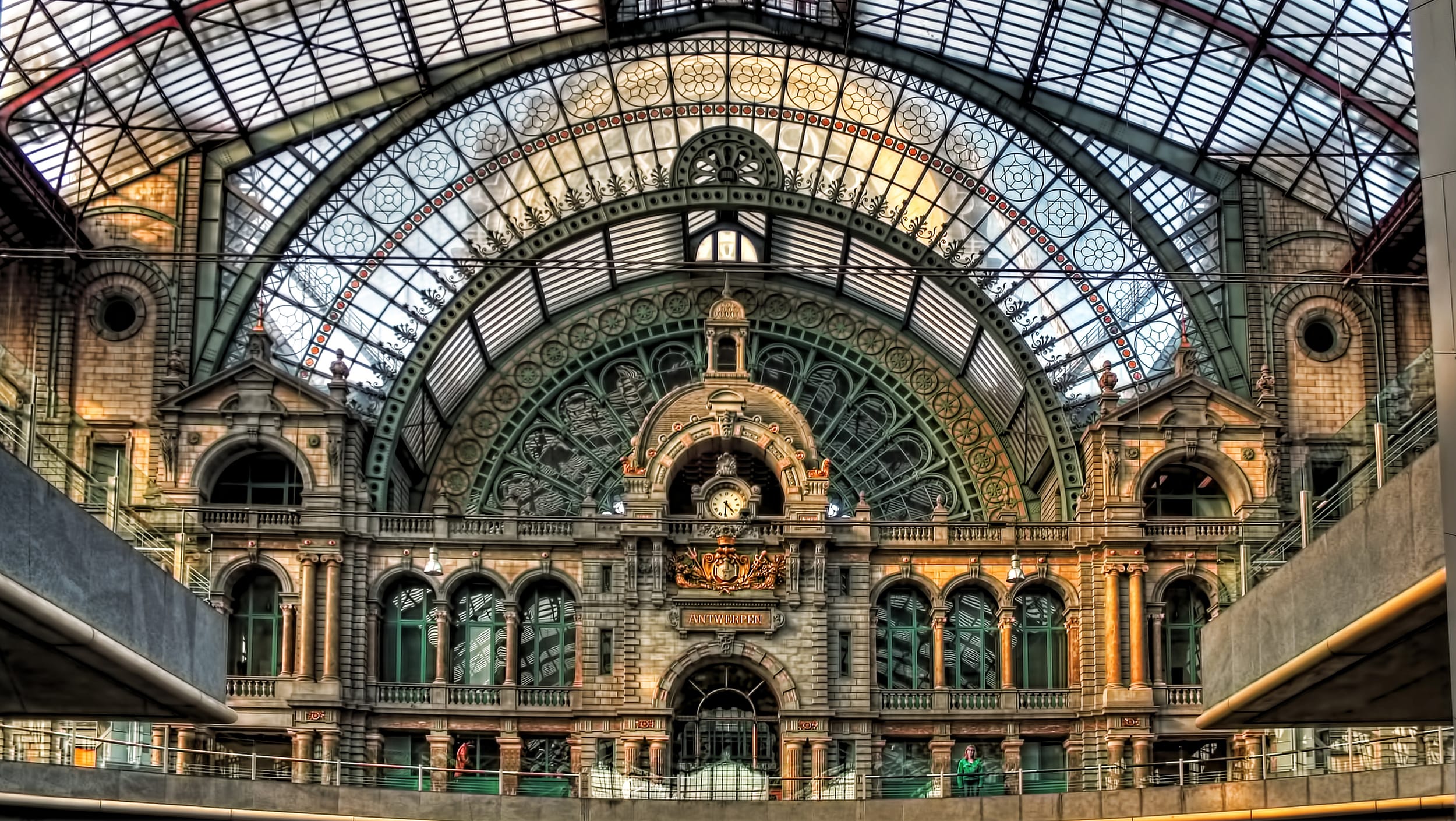 Ornate interior of historic train station with arched glass ceiling and intricate architectural details, featuring a central clock.