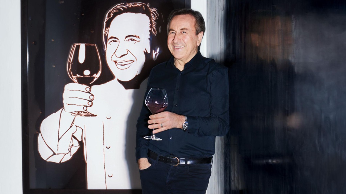 Man in a black shirt holding a wine glass, standing beside a painting of a smiling person holding a glass of wine.