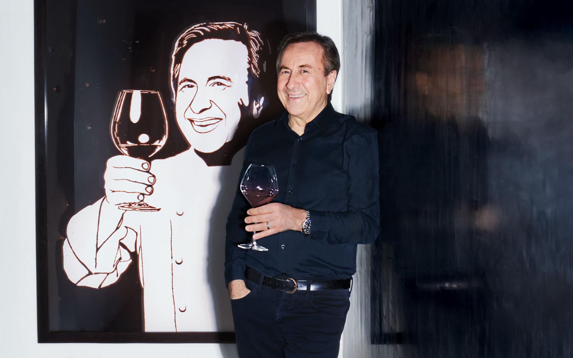 Man in a black shirt holding a wine glass, standing beside a painting of a smiling person holding a glass of wine.