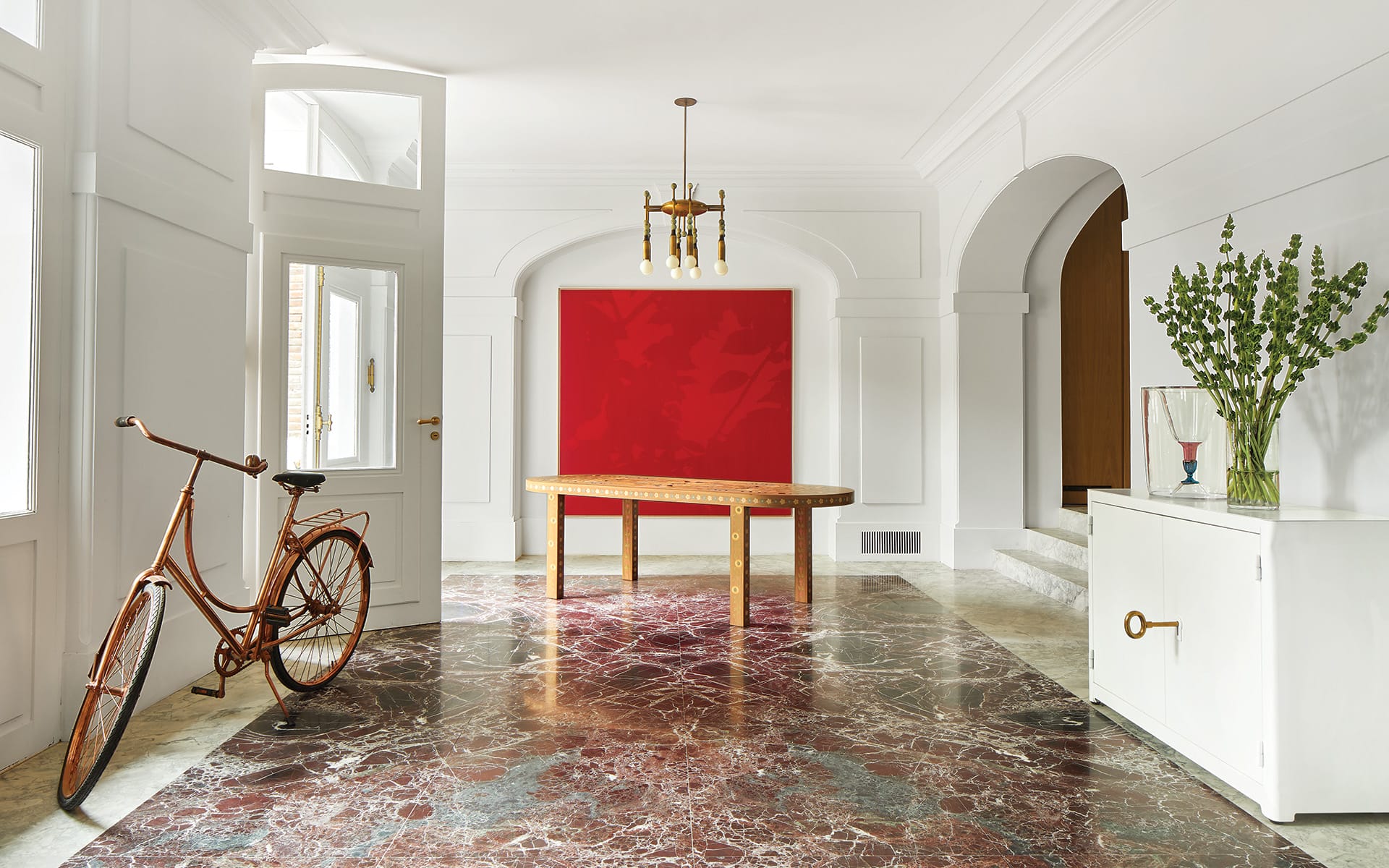 Elegant hallway with red marble floor, vintage bicycle, modern art, and a decorative table under a chandelier.