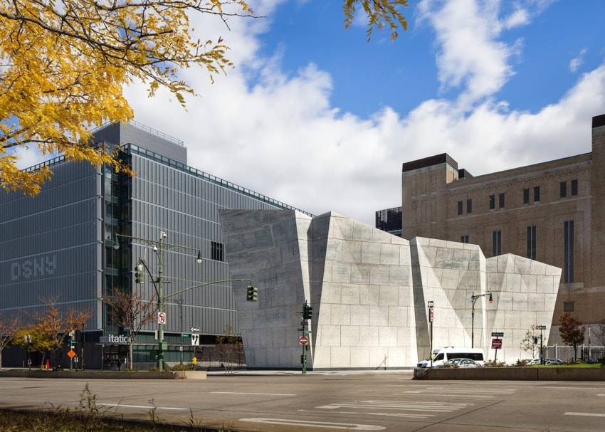 Modern building with angular design and glass facade beside a brick building on a city street under a blue sky.
