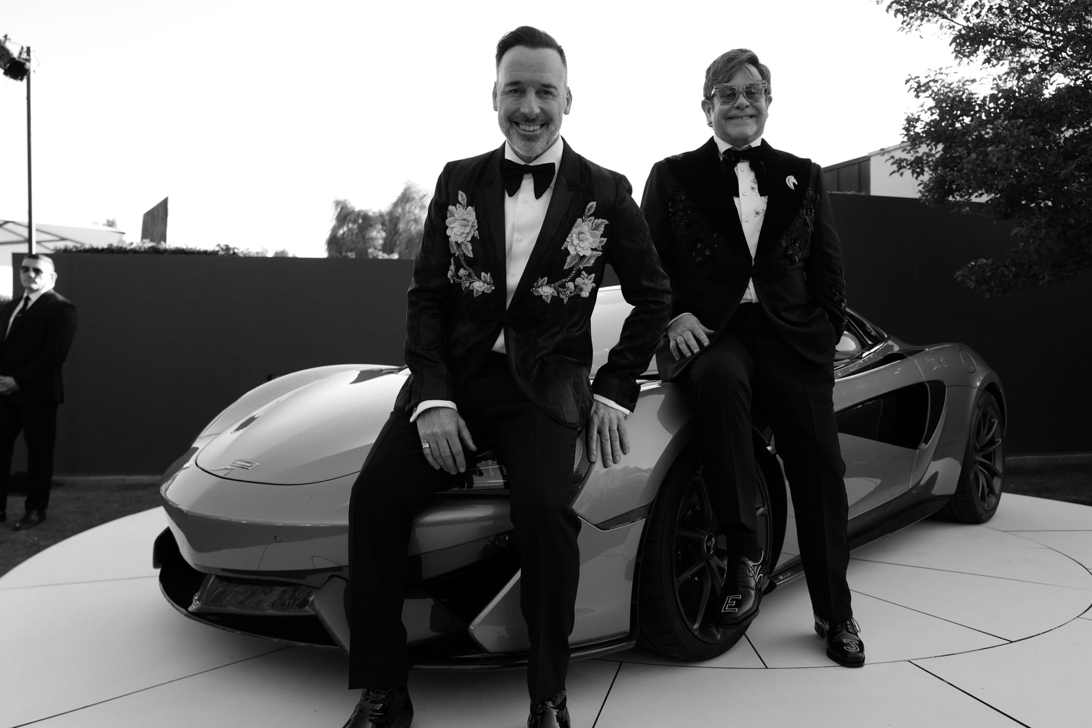 Two men in tuxedos sitting on a sleek sports car at an outdoor event, black and white image.