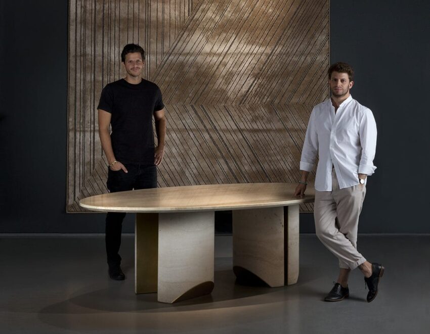 Two men standing beside a modern oval table with a textured wall art piece in the background.