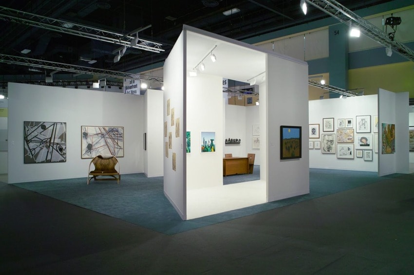 Art exhibition with various paintings on white walls, a chair, and a bench in a spacious gallery setting.