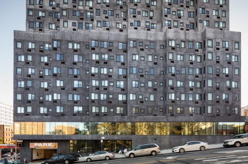 Modern multi-story concrete building with numerous windows on an inclined street, featuring a parking lot at street level.
