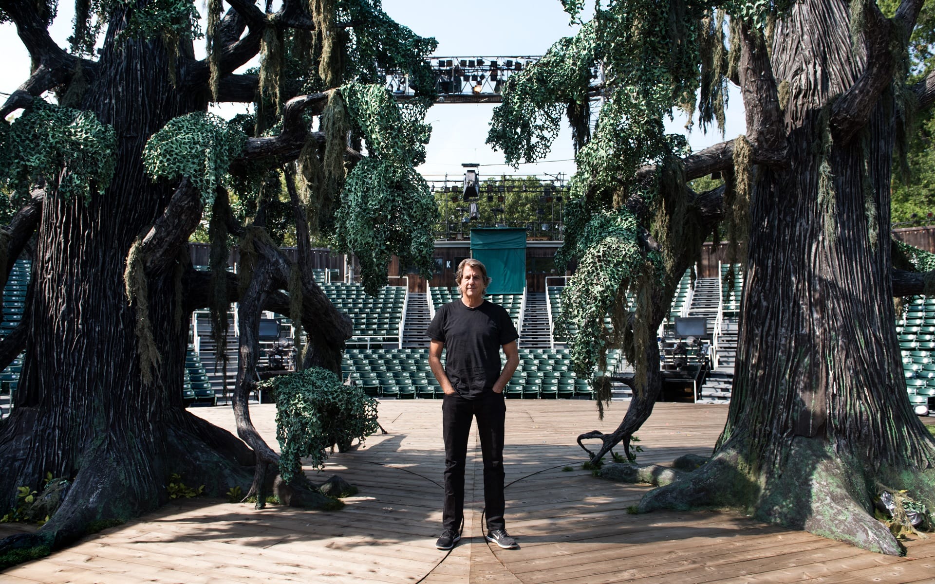 A person standing on an outdoor stage surrounded by large, decorative trees with empty seating in the background.