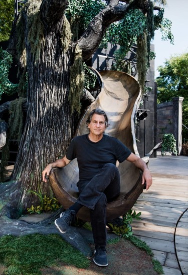 Man sitting on a tree-themed outdoor set, relaxed and casual, with lush greenery and wooden textures in the background.