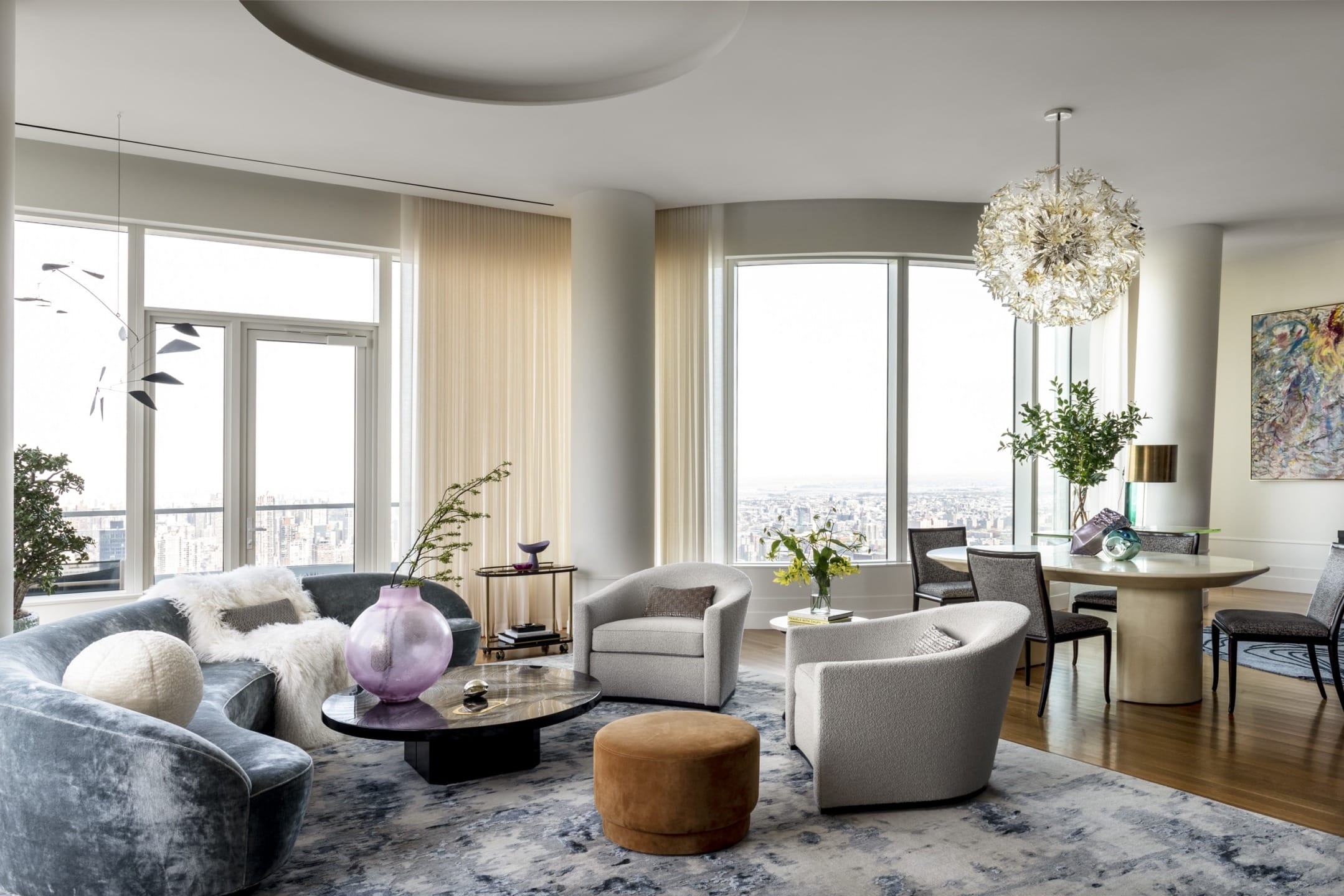 Modern living room with large windows, stylish furniture, round table, unique chandelier, city view, and decorative plants.