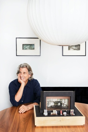 Smiling person sitting at a table with artworks and a large round lamp overhead.