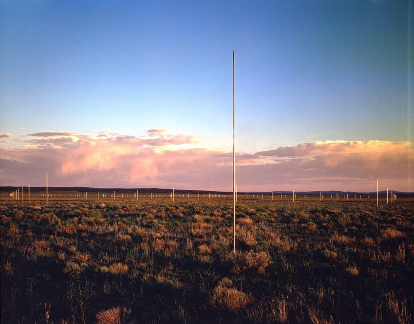 Vast field at sunset with tall vertical poles scattered across the grassy landscape under a dramatic sky.