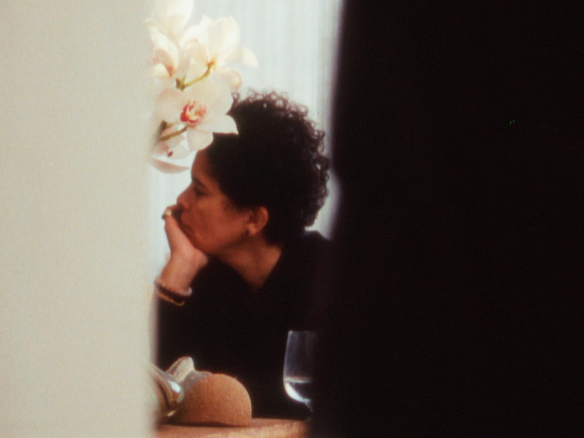 Person with curly hair sits at a table, chin resting on hand, with a white orchid and a glass in the background.