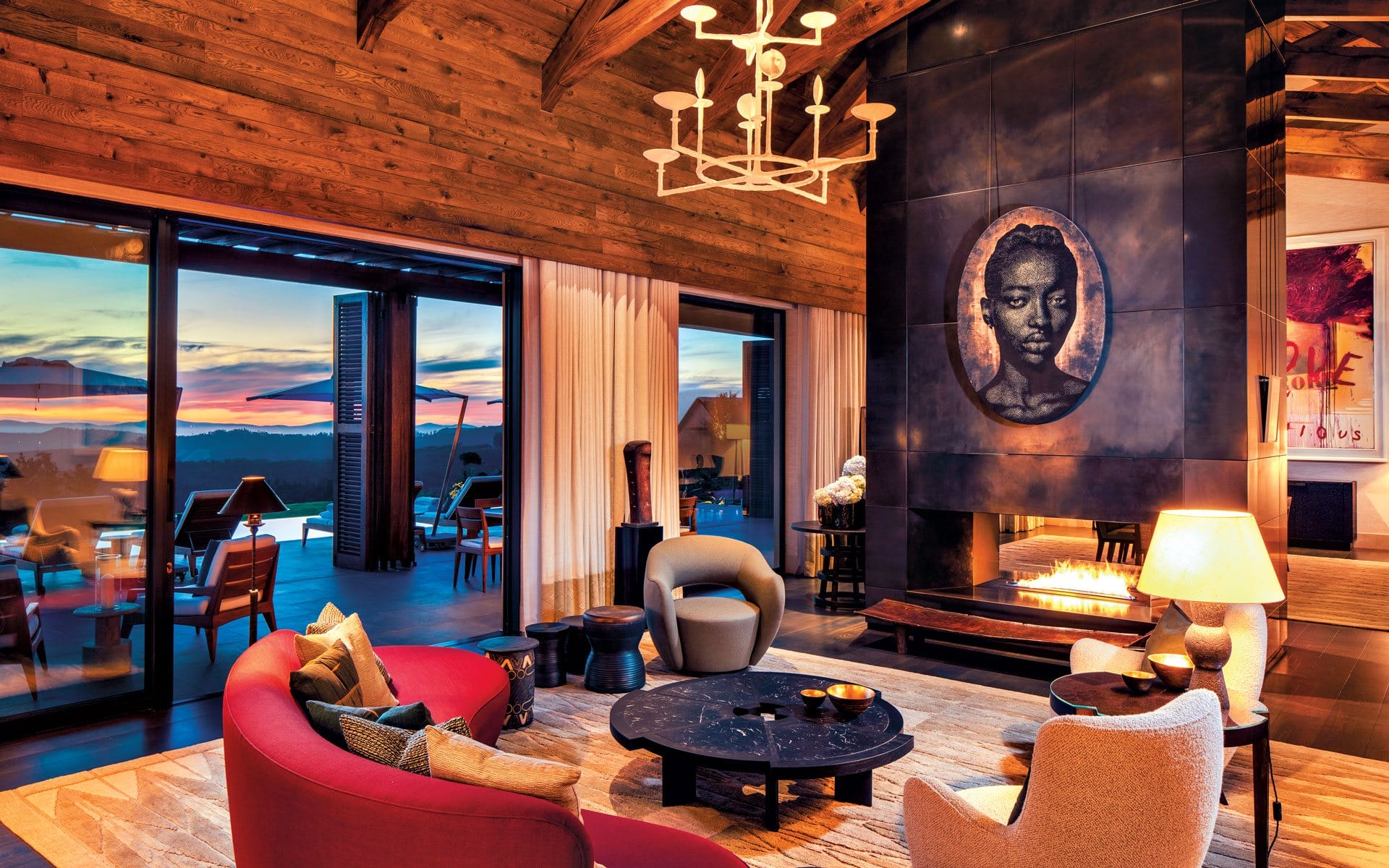 Luxurious living room with modern decor, large windows showing a sunset view, and a prominent portrait above a fireplace.