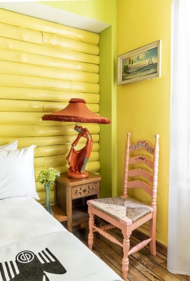 Colorful room with yellow walls, unique orange lamp, pink chair, and small wooden table beside a bed with white linens.
