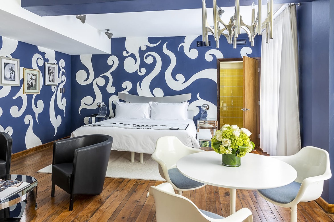 Hotel room with modern blue and white decor, wooden flooring, bed with pillows, small dining table, chairs, and wall art.