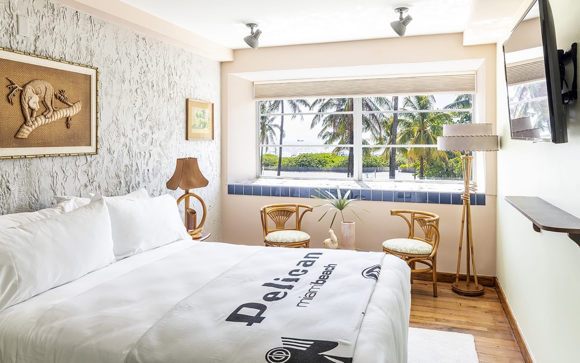 Bright bedroom with white bedding, tropical decor, large window view of palm trees and beach, wicker chairs, and wooden floor.