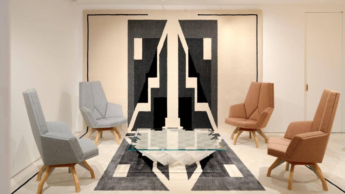Modern lounge with geometric black-and-white rug, glass table, and four angled armchairs in a bright, minimalist space.
