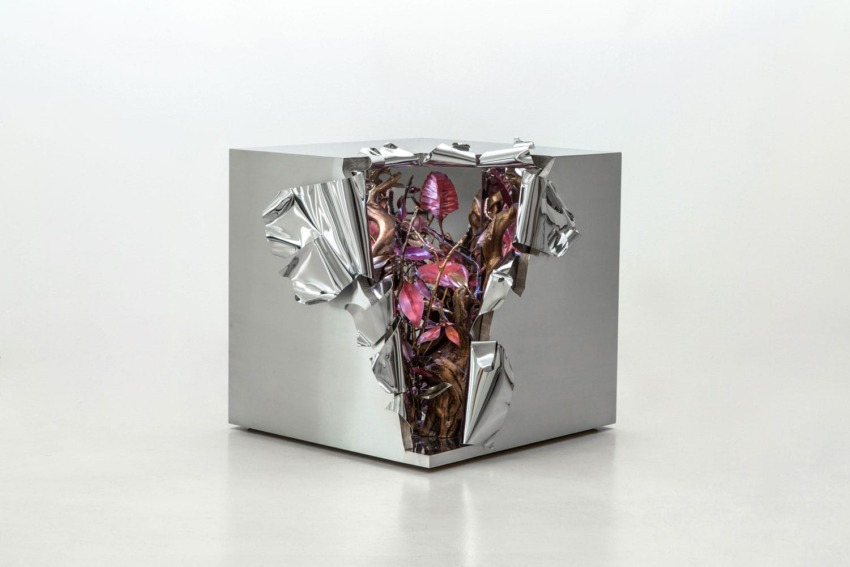 Silver cube sculpture with jagged edges revealing bright pink and purple organic shapes inside, displayed on a light background.