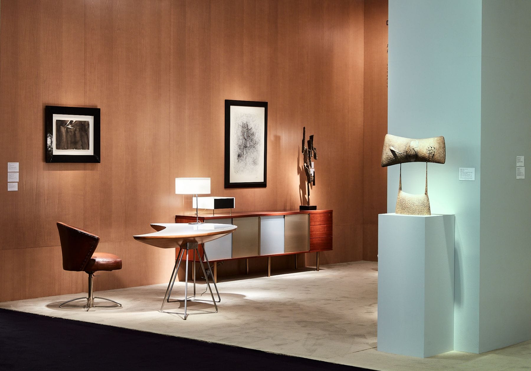 Modern art gallery with sculptures, framed artworks on wooden walls, and contemporary furniture including a chair and table.