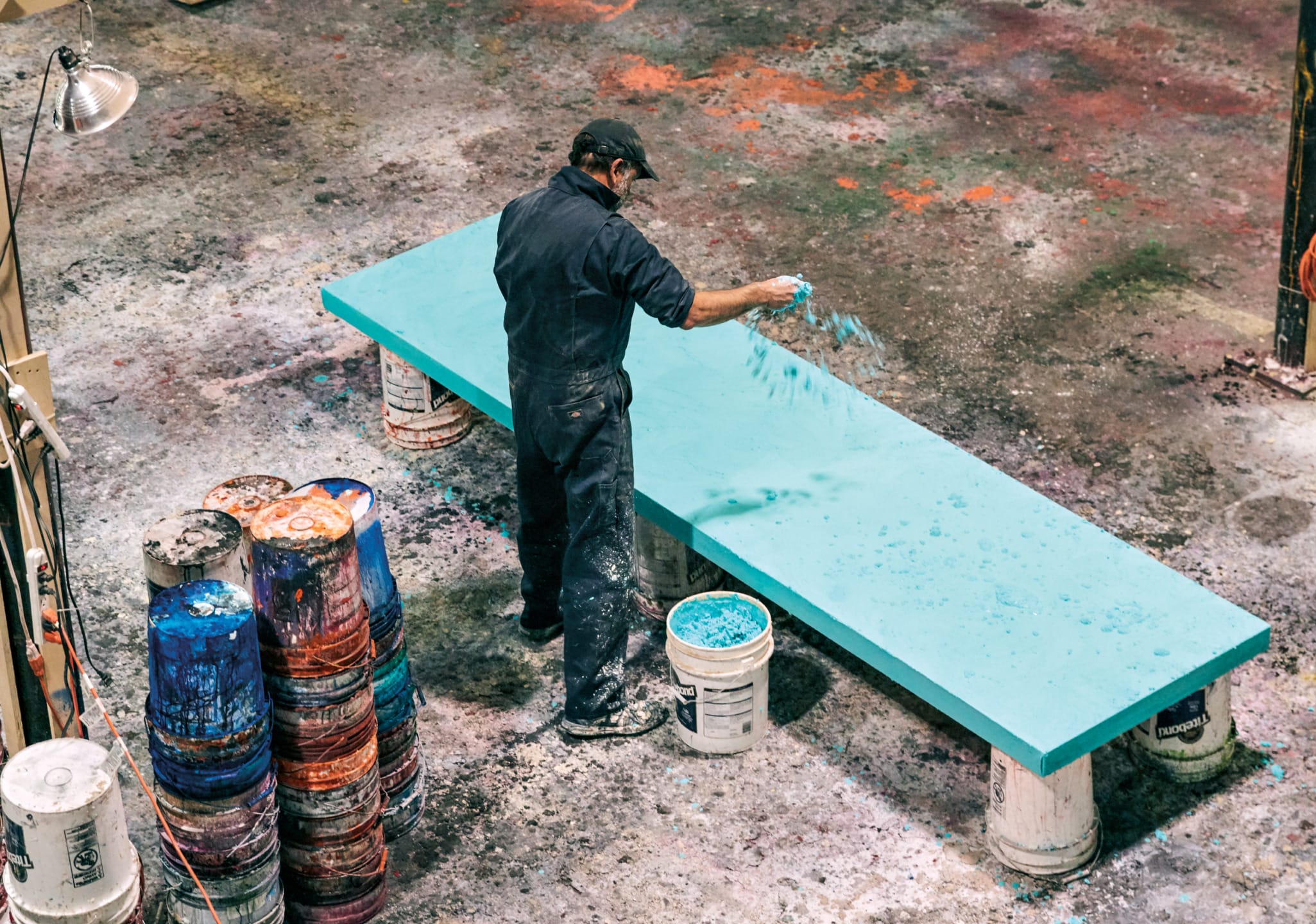 Artist applying blue paint to large canvas in colorful studio setting, surrounded by paint buckets and vibrant floor stains.