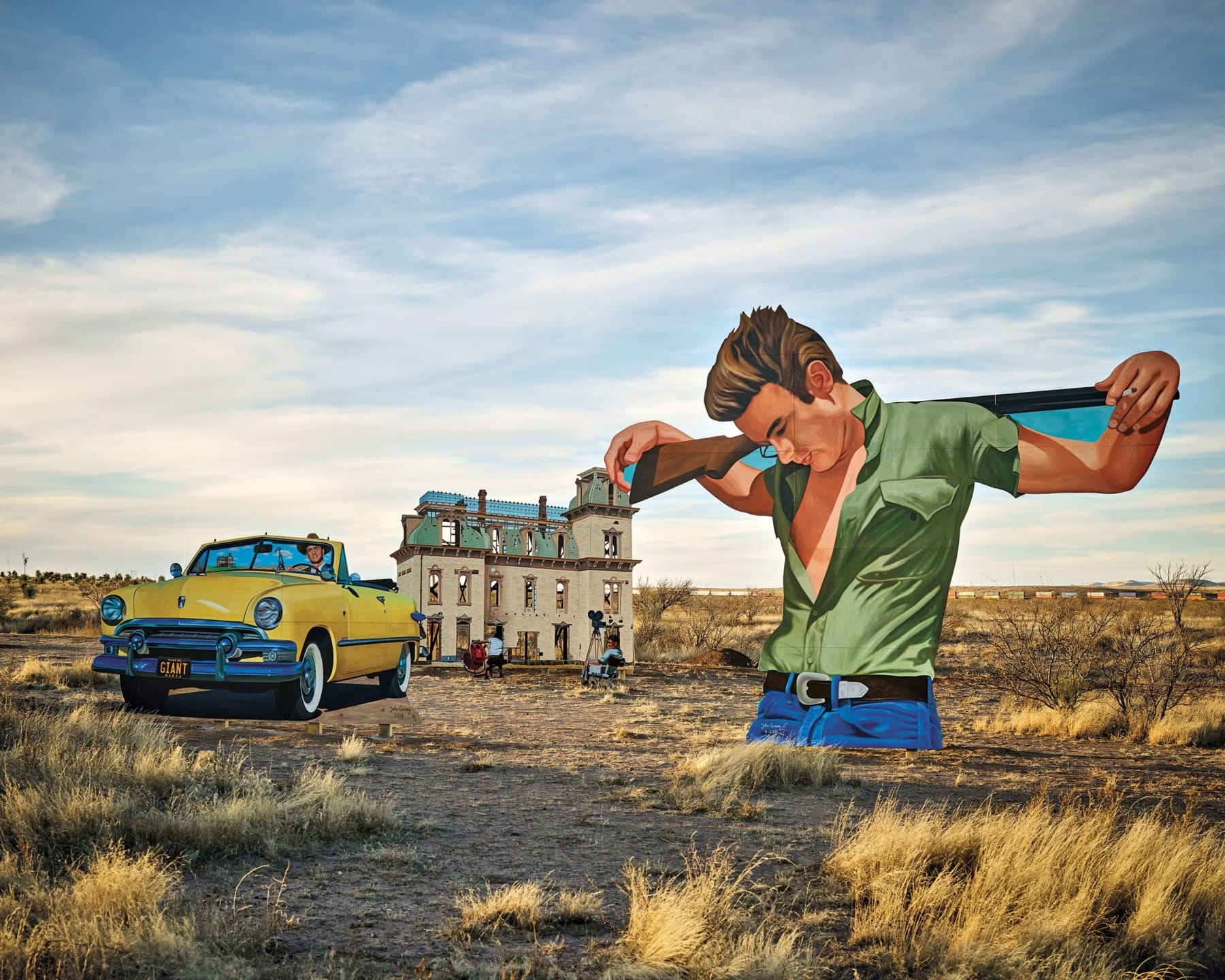 Vintage car and large man mural near an abandoned house in a dry, grassy landscape under a cloudy sky.