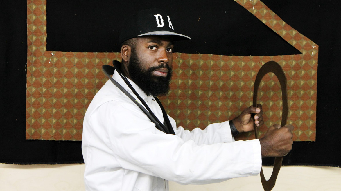 Man in a white coat and cap holding a circular frame against a geometric patterned backdrop.